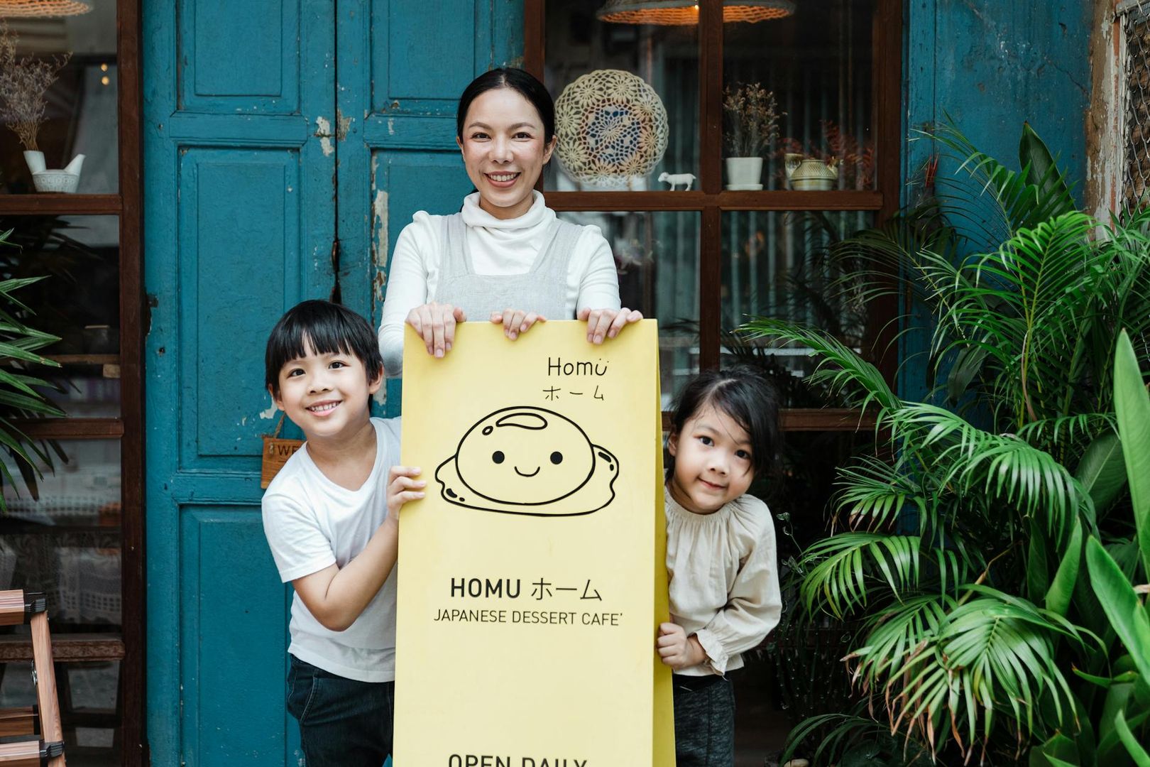 Vrouw en twee lachende kinderen tonen een geel bord van 'Homu Japanese Dessert Café'.